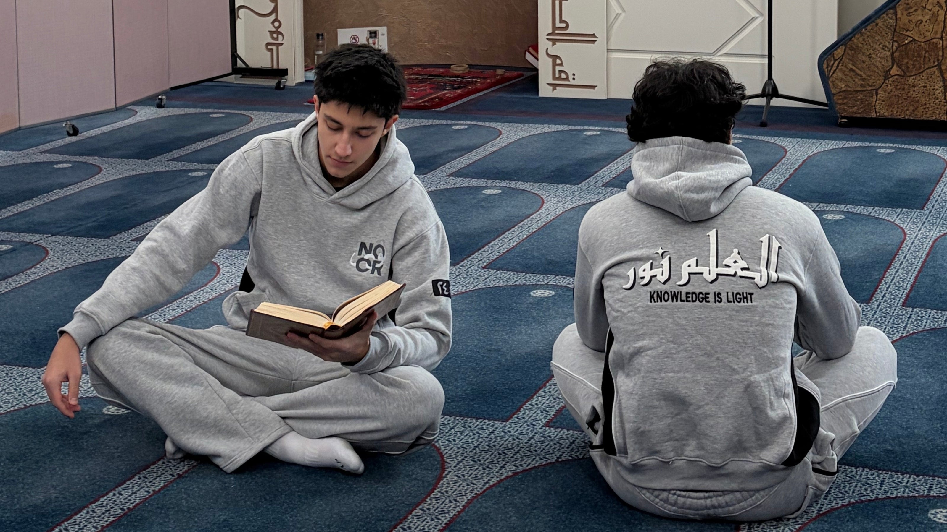 Two people in gray sweatshirts sitting on a blue prayer mat with Arabic text on a white wall.