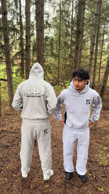 Two people wearing matching gray hoodies and pants in a forest setting. Gray Muslim streetwear brand low cost from Noor Apparel.