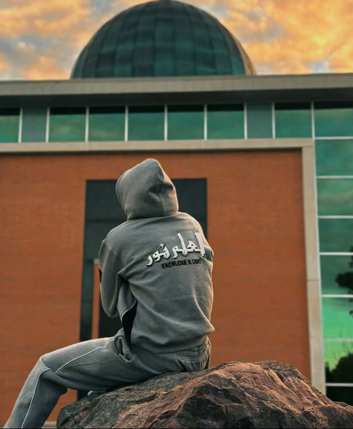 Person wearing a gray hoodie with text sitting on a rock in front of a mosque with a Noor Apparel Hoodie.