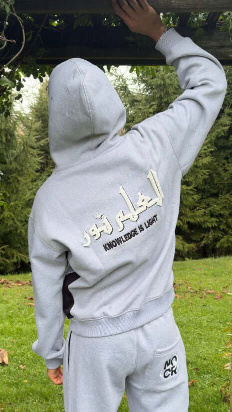 Person wearing a gray hoodie with Arabic text and 'Knowledge is Light' in English, standing outdoors. Noor Apparel Model.