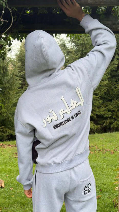 Person wearing a gray hoodie with Arabic text and 'Knowledge is Light' in English, standing outdoors. Noor Apparel Model.